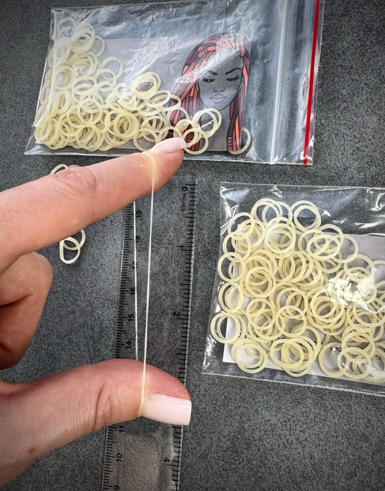 May include: Two clear plastic bags filled with small, off-white elastic hair bands. One bag features a graphic illustration of a person with braided hair. A clear plastic ruler shows one elastic band stretched to approximately 2.5 inches in length.