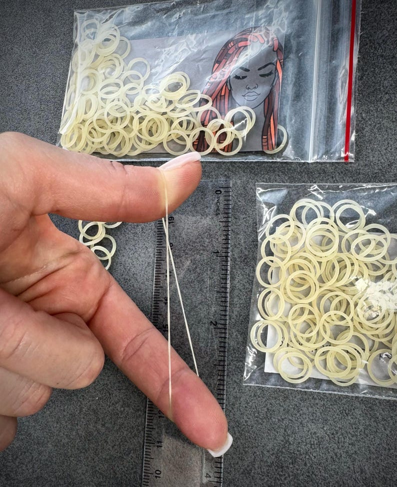 May include: Two clear plastic bags filled with small, translucent off-white elastic hair bands. A transparent plastic ruler is positioned vertically, showing the bands have a diameter of approximately 0.5 inches when stretched between fingers.