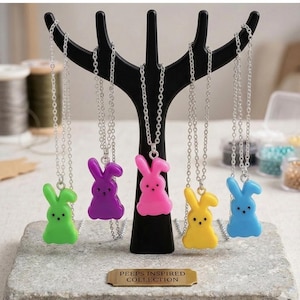 May include: A display of five colorful bunny pendant necklaces. The pendants are green, purple, pink, yellow, and blue. The necklaces hang from a black tree-shaped display stand. The text on the display reads "PEEPS INSPIRED COLLECTION."