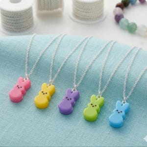 May include: A collection of five colorful bunny-shaped pendant necklaces. Each necklace features a different pastel color: pink, yellow, purple, green, and blue. The pendants are attached to delicate silver chains. The necklaces are displayed on a light blue fabric.