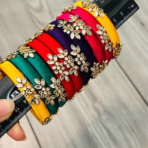 May include: A set of seven multi-colored silk thread bangles in yellow, green, red, and purple, each decorated with a gold-tone floral motif featuring clear crystals. A black ruler shows the stack measures approximately 2.5 inches in diameter.