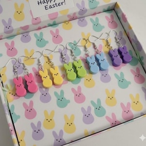 Könnte beinhalten: Ohrringe in Form von bunten Osterhasen in Rosa, Gelb, Grün, Blau und Lila. Die Ohrringe werden in einer Schachtel mit Hasenmuster und einer Karte mit der Aufschrift "Happy Easter!" präsentiert.