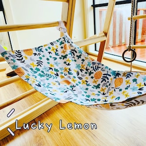 May include: A white fabric hammock with a floral and lemon pattern is suspended from a wooden climbing structure. The pattern includes yellow, blue, and green flowers and lemons. The text "Lucky Lemon" is visible at the bottom.