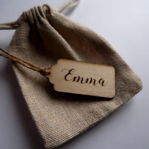 May include: A small, natural-coloured hessian bag with a wooden gift tag attached. The tag is inscribed with the name "Emma" in cursive.