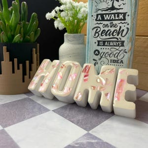 May include: White ceramic "HOME" sign with pink and white shell accents. The sign is sitting on a black and white chequered surface.