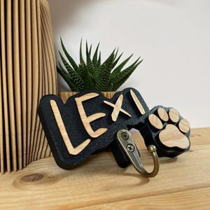 Puede incluir: Un colgador de pared de madera negra con el nombre "LEXI" en madera clara, con un diseño de huella de pata. El gancho es de metal y está fijado al letrero de madera. Una pequeña planta suculenta está en el fondo.