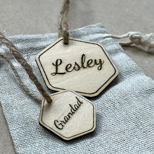 Può includere: Due etichette esagonali in legno con i nomi "Lesley" e "Grandad" incisi in corsivo. Le etichette sono attaccate a un sacchetto con coulisse in fibra naturale. Le etichette sono in legno chiaro.