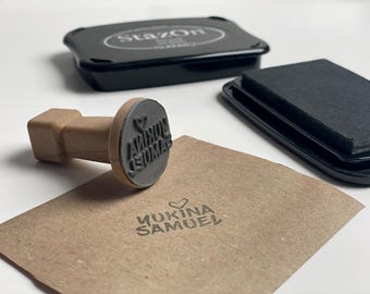 Könnte beinhalten: Ein Holzstempel mit dem Text "AMINON EDOMA" in einem kreisförmigen Design, neben einem schwarzen Stempelkissen und einem gestempelten braunen Papier mit dem Text "YUKINA SAMUEL". Das Stempelkissen ist schwarz und der Stempel ist braun.