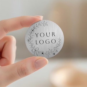 May include: A circular, brushed metal product featuring the text "YOUR LOGO" within a floral wreath. The product is held between two fingers. The design is simple and elegant, suitable for branding or promotional use.