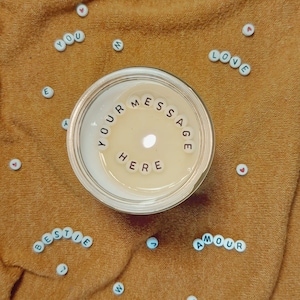 May include: A lit candle in a clear glass container with the words "YOUR MESSAGE HERE" spelled out in small white beads. Scattered around the candle are more beads spelling out words like "LOVE", "BESTIE", and "AMOUR". The candle sits on a textured, orange surface.