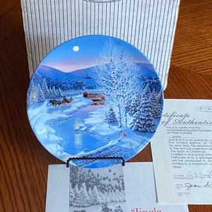 May include: A decorative plate with a winter scene, featuring a snowy landscape, a river, and a horse-drawn carriage. The plate is displayed with a certificate of authenticity and a box. The plate is from the "Jingle Bells" series.