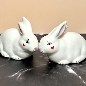 May include: Two white ceramic bunny figurines. Each bunny has black eyelashes, a red heart on its cheek, and pink accents on the ears and nose. The bunnies are positioned close together, appearing to touch noses.