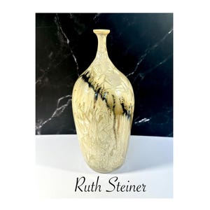 May include: A tall, cream-colored ceramic vase with a textured surface and a narrow neck. The vase features a pattern of etched lines and dark brown accents. The name "Ruth Steiner" is printed at the bottom.