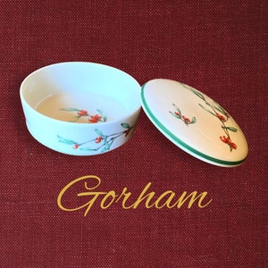 May include: A round white porcelain trinket box with a matching lid, featuring a painted pattern of green holly leaves and red berries. The word Gorham is printed in yellow script below the container. The box has a smooth finish and a green rim.