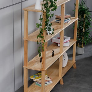 Op de afbeelding: Een licht houten boekenkast met vijf planken. De planken zijn gevuld met boeken, een kleine plant en decoratieve items. De boekenkast heeft een eenvoudig ontwerp met afgeronde randen en poten.