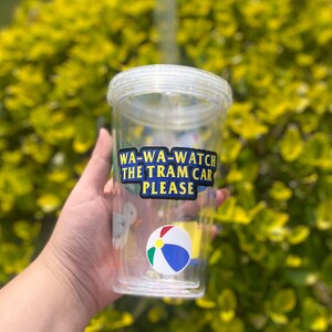 以下が含まれることがあります： ストローが付いた透明なプラスチック製のタンブラーで、カラフルなステッカーが貼られています。ステッカーには「WA-WA-WATCH THE TRAM CAR PLEASE」と書いてあり、漫画のビーチボールと鳥も描かれています。