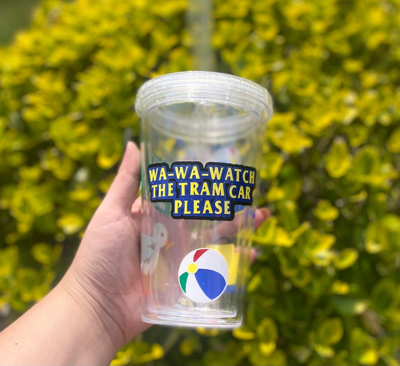 以下が含まれることがあります： ストローが付いた透明なプラスチック製のタンブラーで、カラフルなステッカーが貼られています。ステッカーには「WA-WA-WATCH THE TRAM CAR PLEASE」と書いてあり、漫画のビーチボールと鳥も描かれています。