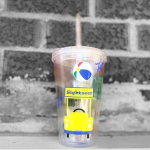 以下が含まれることがあります： ストローが付いた透明なプラスチック製のタンブラー。黄色と青のビーチボール、青色の屋根が付いた黄色の車、そして「Sightseer」という文字が描かれています。