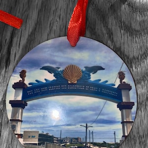 May include: A round, wooden ornament with a colorful image of a seaside archway. The archway is decorated with dolphins and a seashell. The text on the archway reads "You are now leaving the boardwalk of fame & happiness".