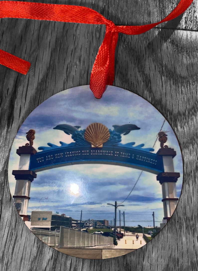 May include: A round, wooden ornament with a colorful image of a seaside archway. The archway is decorated with dolphins and a seashell. The text on the archway reads "You are now leaving the boardwalk of fame & happiness".