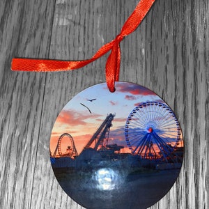 May include: Round ornament with a photo of a Ferris wheel and roller coaster at sunset. The ornament is red and has a red ribbon attached.