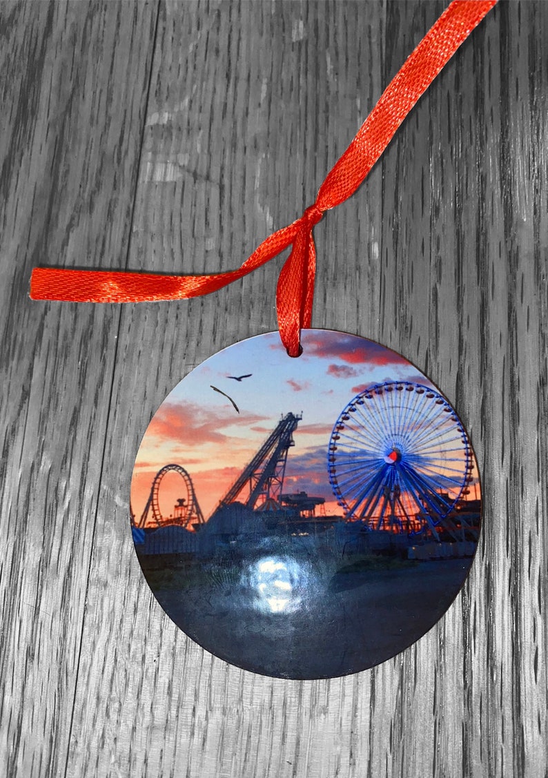May include: Round ornament with a photo of a Ferris wheel and roller coaster at sunset. The ornament is red and has a red ribbon attached.