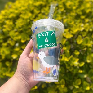 以下が含まれることがあります： ストローが付いた透明なプラスチック製のタンブラー。カモメのグラフィックと、「出口4 ワイルドウッド」と書かれた緑と白の標識が描かれています。