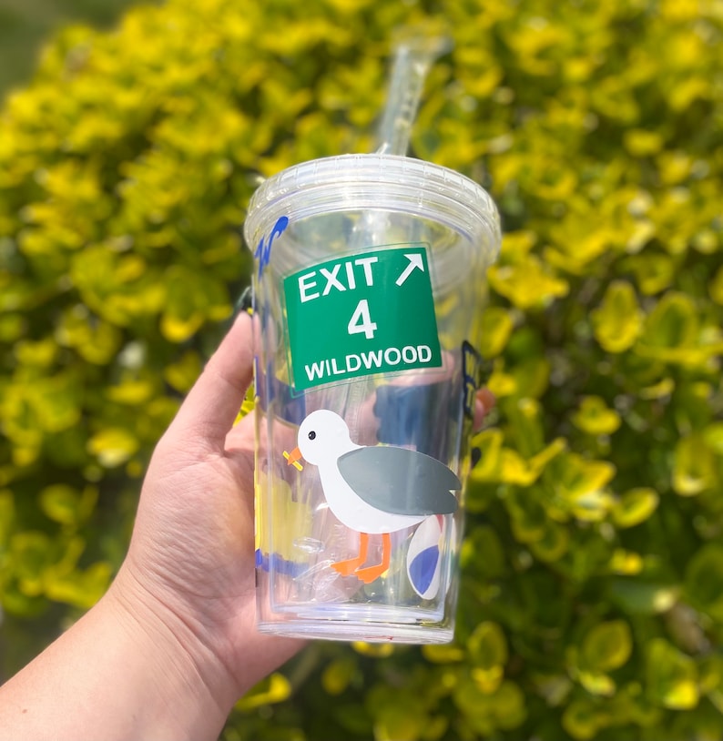 以下が含まれることがあります： ストローが付いた透明なプラスチック製のタンブラー。カモメのグラフィックと、「出口4 ワイルドウッド」と書かれた緑と白の標識が描かれています。
