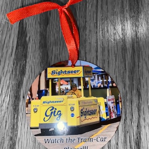 May include: A yellow and blue tram car with the words "Sightseer Gig" on the side. The tram car is decorated with red, white, and blue streamers. The tram car is on a wooden surface with a red ribbon attached to it. The text "Watch the Tram-Car Please!!!" is printed below the tram car.