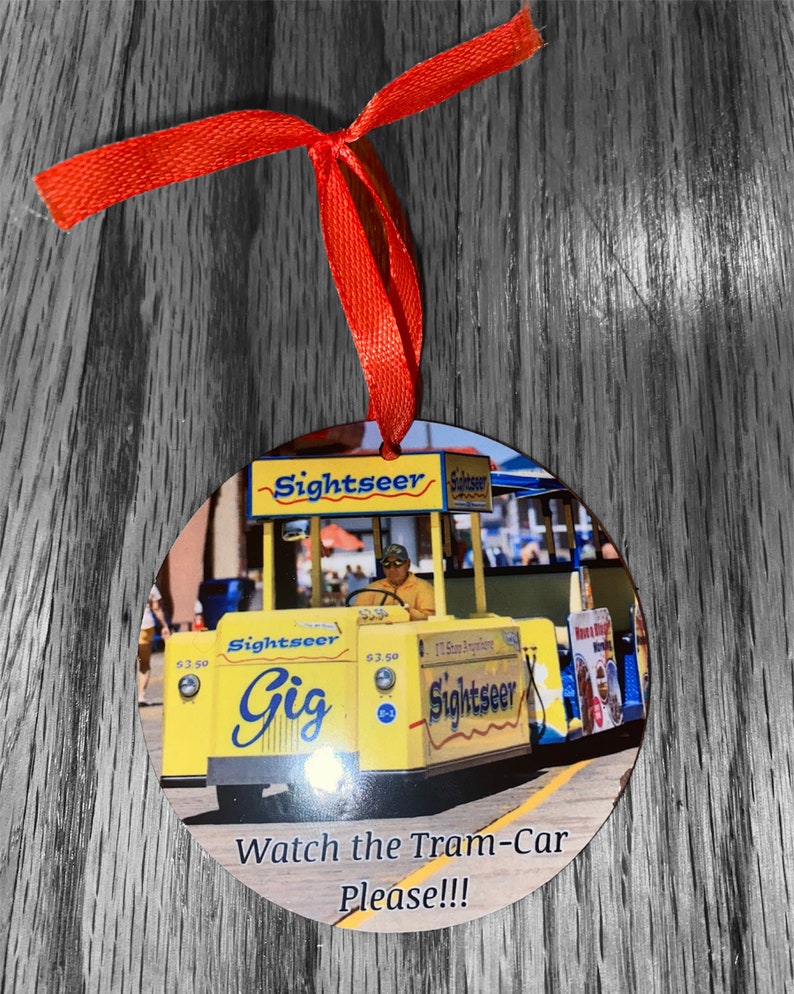 May include: A yellow and blue tram car with the words "Sightseer Gig" on the side. The tram car is decorated with red, white, and blue streamers. The tram car is on a wooden surface with a red ribbon attached to it. The text "Watch the Tram-Car Please!!!" is printed below the tram car.