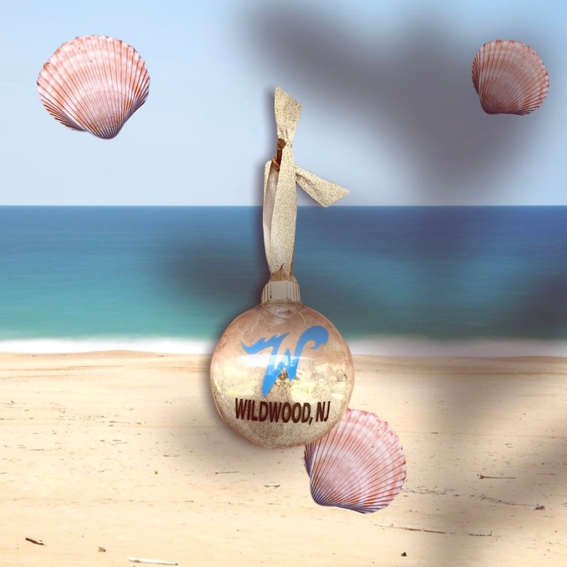 May include: A clear glass ornament with gold glitter inside and a blue and white graphic of a wave and the text "Wildwood, NJ". The ornament is hanging from a gold ribbon and there are three seashells in the background.
