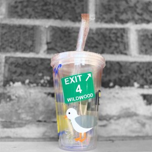 以下が含まれることがあります： 緑と白の出口標識のステッカーが貼られた透明なプラスチック製のタンブラーで、「Exit 4 Wildwood」と書いてあります。タンブラーにはストローと、漫画の海鳥のイラストが描かれています。