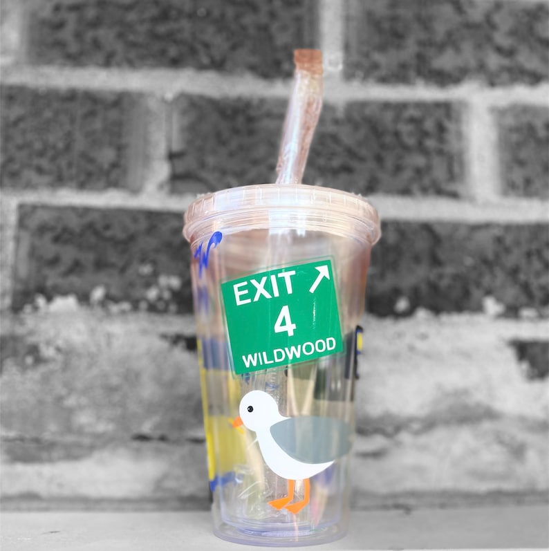 以下が含まれることがあります： 緑と白の出口標識のステッカーが貼られた透明なプラスチック製のタンブラーで、「Exit 4 Wildwood」と書いてあります。タンブラーにはストローと、漫画の海鳥のイラストが描かれています。