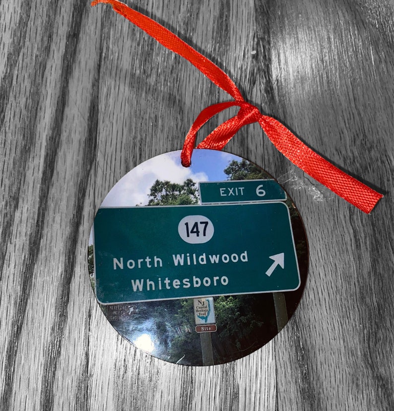 May include: A round, white, and green Christmas ornament with a photo of a highway sign. The sign reads "Exit 6", "147", "North Wildwood", and "Whitesboro". An arrow points to the right.