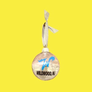 May include: A clear glass Christmas ornament with a silver glitter finish. The ornament has a blue "W" with a white outline and the text "WILDWOOD, NJ" printed on it. The ornament has a silver ribbon attached to the top.