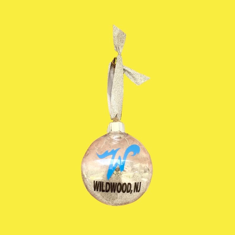 May include: A clear glass Christmas ornament with a silver glitter finish. The ornament has a blue "W" with a white outline and the text "WILDWOOD, NJ" printed on it. The ornament has a silver ribbon attached to the top.