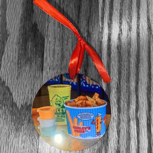 May include: A round, white ornament with a picture of a yellow and blue container of curly fries. The container has the text "Curly's Fries" and a cartoon character holding a fry. There are three small containers of dipping sauces and a yellow container of lemonade.