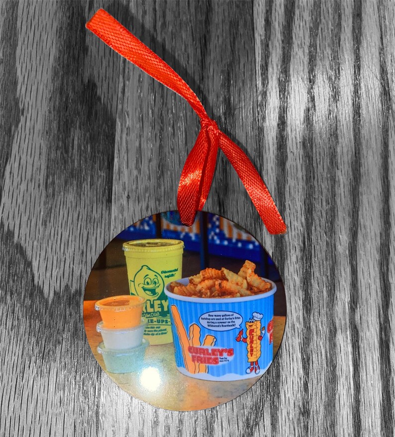 May include: A round, white ornament with a picture of a yellow and blue container of curly fries. The container has the text "Curly's Fries" and a cartoon character holding a fry. There are three small containers of dipping sauces and a yellow container of lemonade.