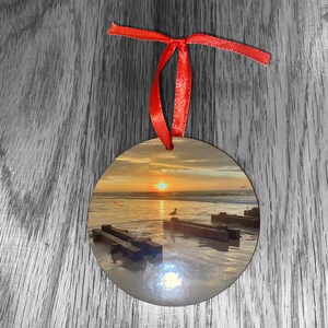 May include: Round wooden ornament with a red ribbon. The ornament features a photograph of a beach scene with a sunset, a seagull, and wooden planks in the water.