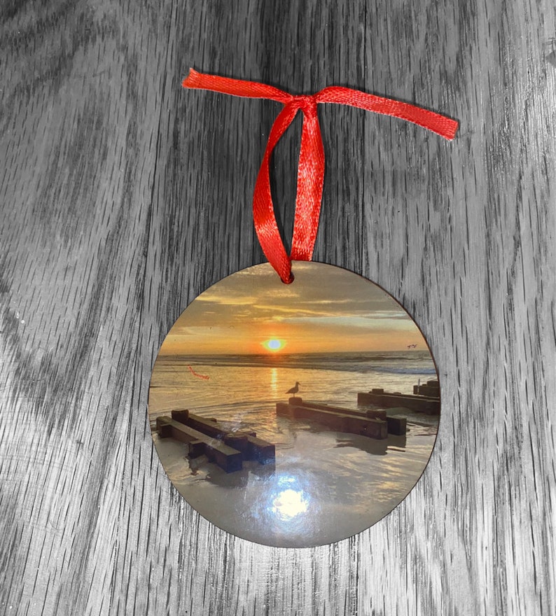 May include: Round wooden ornament with a red ribbon. The ornament features a photograph of a beach scene with a sunset, a seagull, and wooden planks in the water.