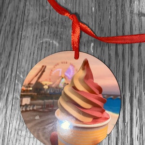 May include: A round wooden ornament with a picture of a pink and yellow swirl ice cream cone in a cup. The background of the ornament is a blurred image of a Ferris wheel and a beach.