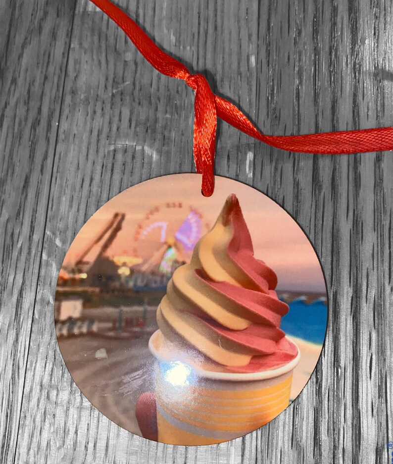 May include: A round wooden ornament with a picture of a pink and yellow swirl ice cream cone in a cup. The background of the ornament is a blurred image of a Ferris wheel and a beach.