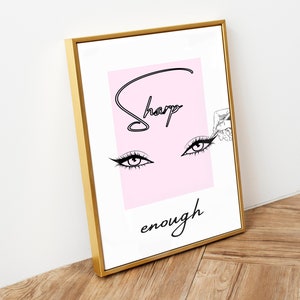 May include: A framed print with a pink background and black line art of eyes with the text "Sharp enough" in black.
