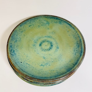 May include: A stack of four ceramic plates with a unique glaze. The plates feature a blend of blue and green hues, with a darker blue rim and a lighter center. The plates have a rustic, handmade appearance.