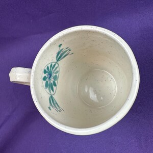 May include: Cream-colored ceramic mug with a speckled finish and a geometric handle. The interior features a teal floral design. The mug is set against a purple background.