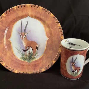 May include: A brown and gold ceramic plate with a black and white border features a painted image of a gazelle. A matching brown and gold ceramic mug with a black and white border features a painted image of a gazelle and a bird.