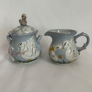 May include: A blue and white porcelain sugar bowl and creamer set with swan and chick designs. The lid of the sugar bowl features a small bird finial.