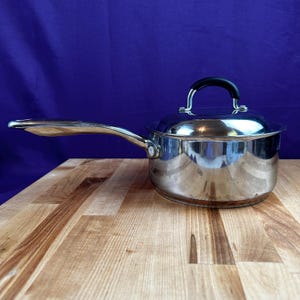 May include: A stainless steel saucepan with a long handle and a lid with a black handle. The pot is sitting on a wooden surface with a blue background. The pot is reflective.