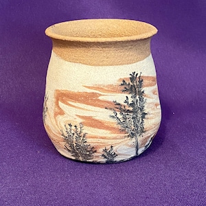 May include: A small, white ceramic vase with a brown rim and a brown and white marbled design. The vase has a black silhouette of trees on the front.