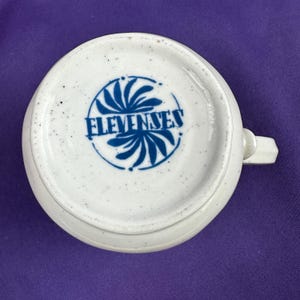 May include: A white ceramic teacup with a handle, viewed from the bottom. The base features a blue circular design with the word "ELEVENESES" in blue lettering. The cup has a speckled texture.